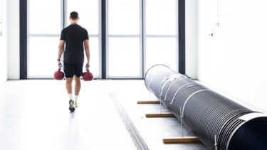 En mann går med en kettlebell i hver hånd med ryggen mot kamera