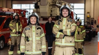 En kvinnelig og en mannlig brannkonstabel smiler mot kamera. I bakgrunnen står tre kolleger.